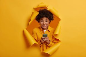 Une personne vêtue d'une chemise jaune sourit en tenant un smartphone et en se tenant dans un trou déchiré sur un fond de papier jaune.