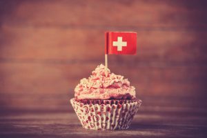 Un cupcake au glaçage rose dans un emballage en forme de cœur est surmonté d'un petit drapeau suisse, sur un fond en bois.
