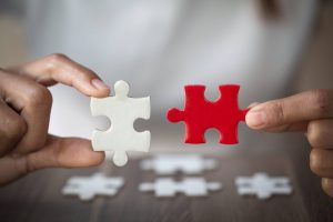 Deux mains tenant des pièces de puzzle, l'une blanche et l'autre rouge, tentant de les assembler au-dessus d'une table contenant d'autres pièces de puzzle.