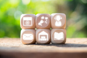 Six blocs de bois présentent des icônes : un globe terrestre, un mégaphone, une personne avec un casque aidant les clients, une enveloppe, une maison et un livre ouvert. Ils sont disposés sur deux rangées sur un fond vert flou.